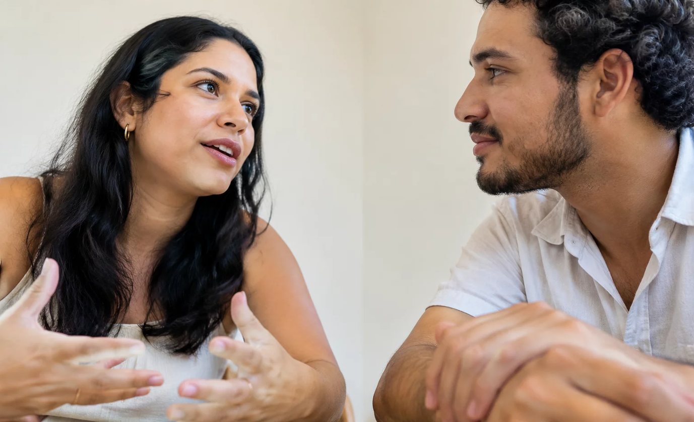 Três participantes conversam ao redor de uma mesa comprida numa sala clara de janelas altas